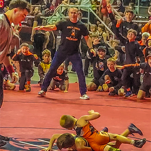 Coach Preston Smith counting back points while watching a wrestling match with a group of youth wrestlers around him